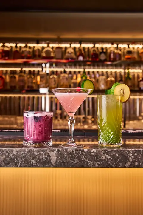 Three cocktails, pink, purple and green, on the counter in front of backlit bottles