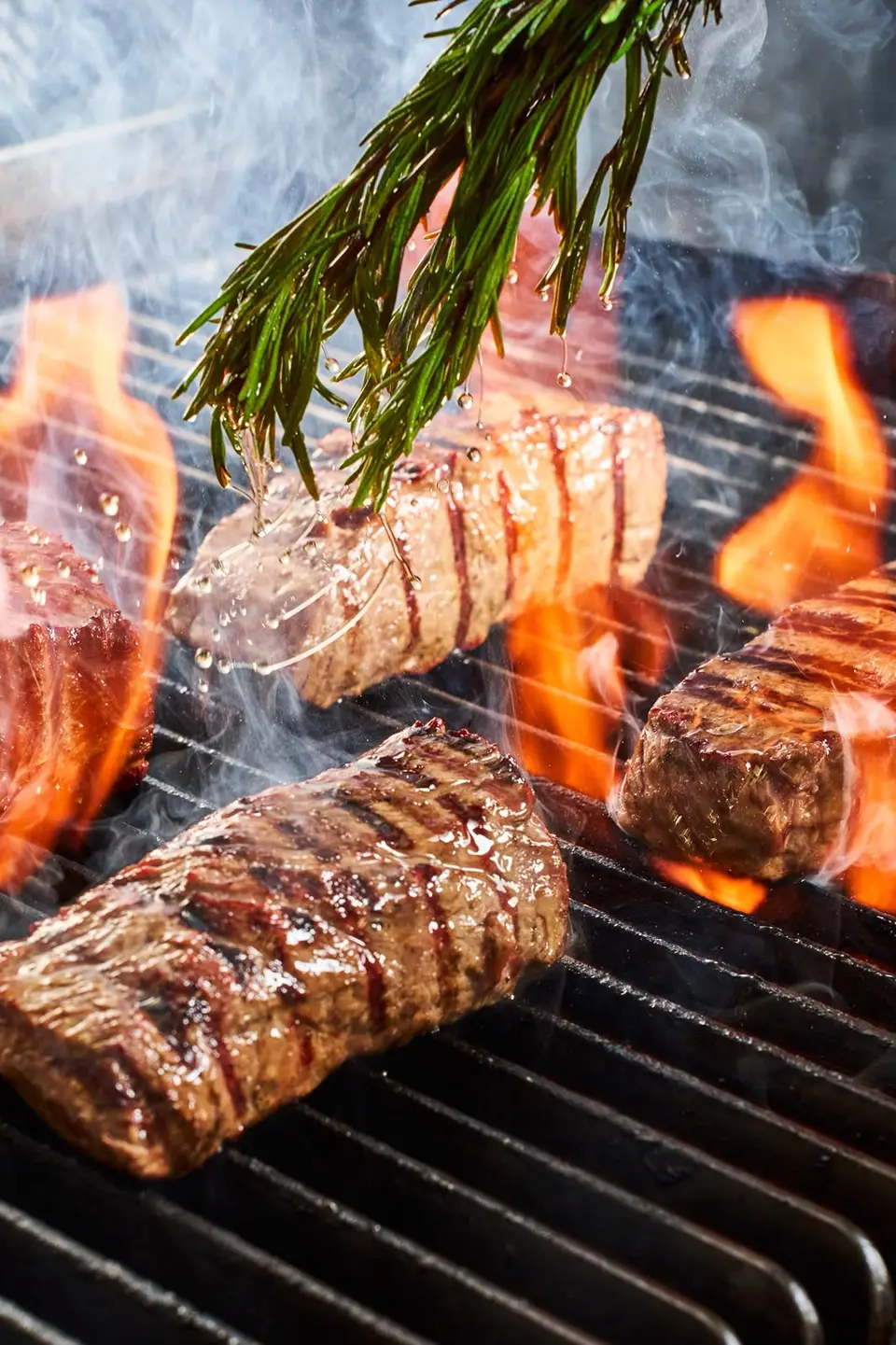 Pièces de bœuf saisies sur le grill au-dessus des flammes, badigeonnées avec un bouquet de romarin
