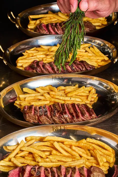 Oval dishes engraved “BEEF” with sliced beef and fries, a hand holding a sprig of rosemary